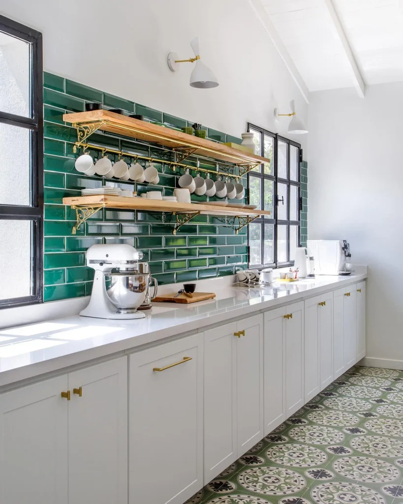Glossy emerald green subway tile wall with light wood shelves on brass brackets and white mugs on a rail