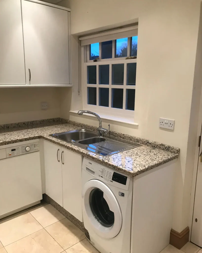 Front loading washer tucked under a granite L-shaped counter with a stainless steel sink beneath a small window and white cabinets