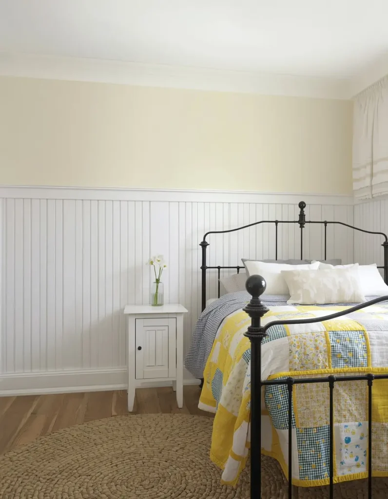 White beadboard half wall under a butter yellow wall with a black iron bed, patchwork quilt and braided jute rug
• 