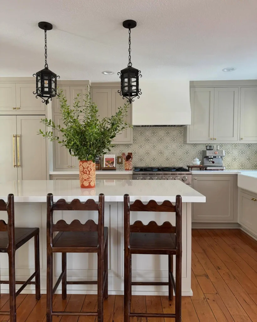Two black wrought iron lanterns over a white island with scalloped wood stools and greige shaker cabinets