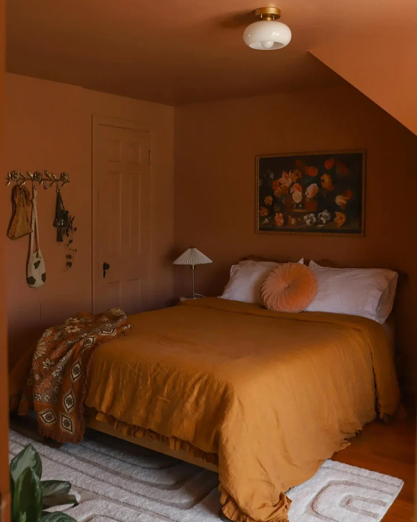 Terracotta painted bedroom with a mustard linen duvet, round velvet pillow, pleated lamp, vintage floral art and a brass hook rail