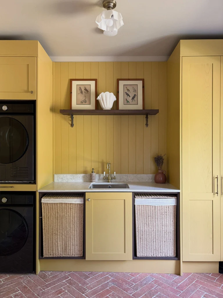 Mustard yellow laundry room with tall cabinets, stacked black washer and dryer, two woven basket cubbies, a small sink, and a brick herringbone floor