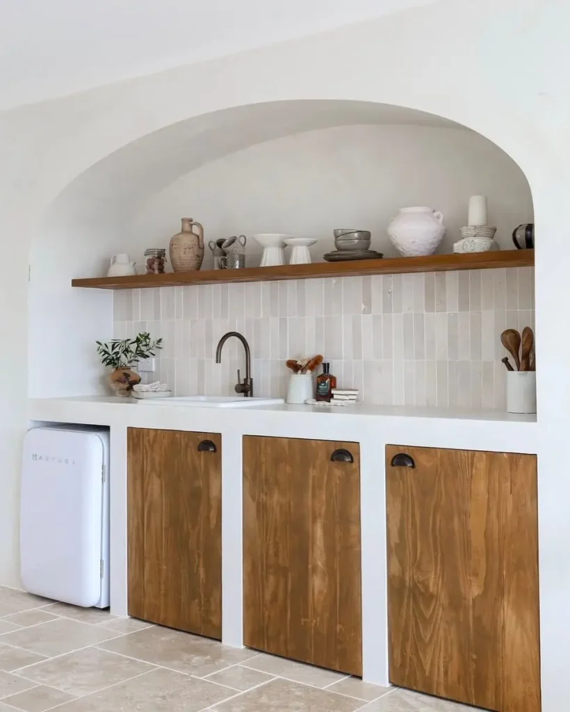Curved white plaster kitchen alcove with a wood shelf of clay jugs, cream vertical tiles, and wood cabinet doors