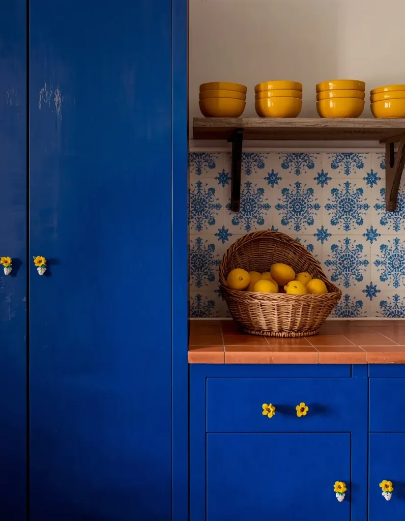 Cobalt blue kitchen cabinets with yellow flower ceramic knobs, a terracotta tile counter, and a basket of lemons