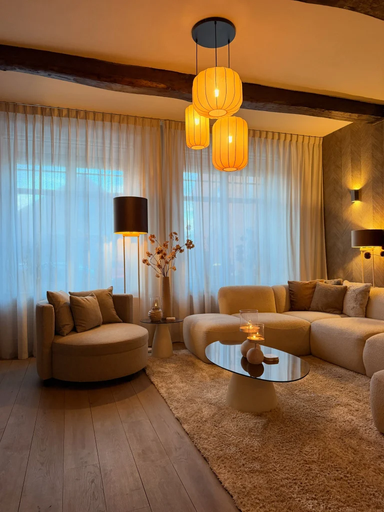 Three orange fabric lantern pendants glowing over a curved bouclé sectional and an oval smoked glass table